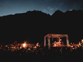 Nächtliche Veranstaltung in den Bergen mit kleiner Bühne, Lagerfeuern und sitzendem Publikum vor dunkler Bergkulisse.