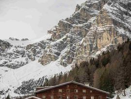 Die Schlickeralm ist im Winter von Schnee bedeckt. Hinter den rustikalen Holzgebäuden erhebt sich eine steile, schneebedeckte Felswand.