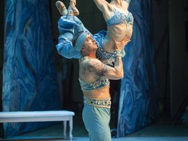 Balletttänzerin in einem blau-türkisen Kostüm wird von einem Tänzer in orientalisch gestaltetem Kostüm während einer Hebefigur auf der Bühne getragen.