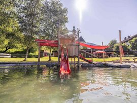 „Kind rutscht auf einer roten Rutsche von einem hölzernen Spielturm in ein flaches Wasserbecken auf einem naturnahen Spielplatz.“