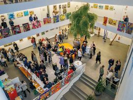 Blick von oben auf die Kunstvernissage im Atrium Amras mit Besuchern, bunten Gemälden an den Galeriegeländern und Sitzbereichen im Innenraum.