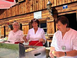 „Drei Frauen bereiten an einem Stand vor einer traditionellen Holzhütte frisch gebackene Krapfen für die Besucherinnen und Besucher zu.“