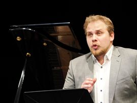 Ein Sänger im hellgrauen Sakko interpretiert Schuberts „Winterreise“ mit konzentriertem Ausdruck vor einem Konzertflügel. Vor ihm steht ein Notenpult.