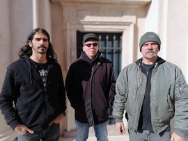 Drei Musiker stehen vor einem Gebäude und präsentieren sich als Band für ein Rock- und Metal-Konzert.