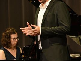 Ein Sänger in schwarzem Anzug interpretiert eine Opernarie ausdrucksstark auf der Bühne, begleitet von einer Pianistin am Flügel.
