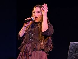 Die Sängerin von Schwarzes Edelweiss singt mit ausdrucksstarker Geste in ein Handmikrofon während eines Bühnenauftritts.