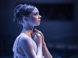 Tänzerin in einem weißen Ballettkostüm steht im blauen Bühnenlicht im Profil und hält die Hände vor dem Oberkörper.