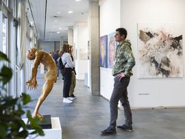 Besucher:innen betrachten Gemälde und eine Skulptur in einem hellen Ausstellungsbereich.