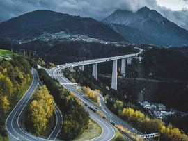 „Luftaufnahme einer kurvenreichen Autobahnbrücke über einem bewaldeten Tal in den Alpen mit Bergpanorama und herbstlicher Landschaft.“