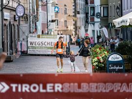 Ein Läufer erreicht in einer Altstadt das Ziel und wird von einer Frau und einem Kind freudig empfangen.