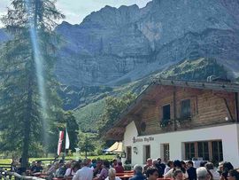 „Besucherinnen und Besucher sitzen bei sonnigem Wetter vor einer Almwirtschaft und genießen Speisen und Getränke vor einer imposanten Bergkulisse.“