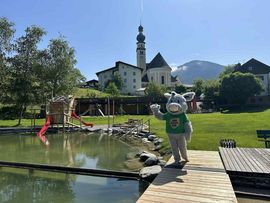„Graues Eselsmaskottchen mit grünem T-Shirt steht auf einer Holzplattform an einem Naturbad mit Spielplatz vor einer Kirche und bewaldetem Bergpanorama.“v