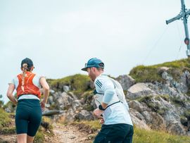 Zwei Trailrunner laufen auf einem felsigen Bergpfad durch eine alpine Landschaft.