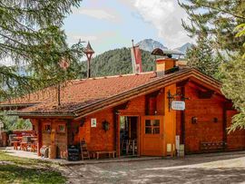 Rustikales Holzgebäude einer Berghütte mit rotem Ziegeldach, umgeben von Nadelbäumen. Im Hintergrund sind bewaldete Berghänge und markante Gipfel zu sehen.