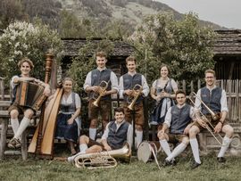Eine Gruppe von acht Musikerinnen und Musikern in traditioneller Tracht posiert vor einem historischen Bauernhof in einer alpinen Landschaft. Zu sehen sind verschiedene Instrumente wie Akkordeon, Harfe, Trompeten, Posaune und Tuba.