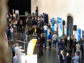 : Besucher:innen und Aussteller bei einer gut besuchten Messe mit zahlreichen Informationsständen.