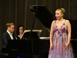 Eine junge Sopranistin singt in einem floralen Abendkleid auf der Bühne, während Pianist und Pianistin sie am Flügel begleiten.
