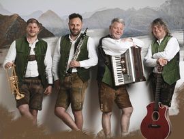 Vier Musiker in traditioneller Tracht stehen gemeinsam vor einer alpinen Bergkulisse. Sie präsentieren verschiedene Instrumente wie Trompete, Klarinette, Akkordeon und Gitarre und vermitteln eine volkstümliche Musikstimmung.