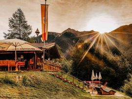 Die Enzianhütte liegt an einem bewaldeten Berghang vor einer eindrucksvollen Bergkulisse. Die tief stehende Sonne taucht die Landschaft in warmes Licht; vor der Hütte befinden sich Sitzplätze, Sonnenschirme und Liegestühle.