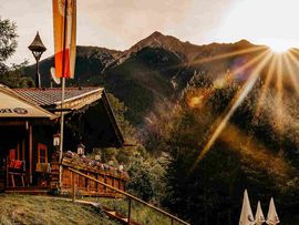 Die Enzianhütte liegt an einem bewaldeten Berghang vor einer eindrucksvollen Bergkulisse. Die tief stehende Sonne taucht die Landschaft in warmes Licht; vor der Hütte befinden sich Sitzplätze, Sonnenschirme und Liegestühle.
