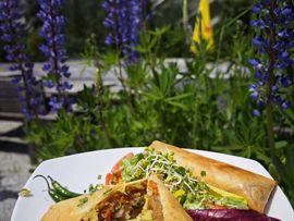 Teller mit mehreren gefüllten Teigtaschen und frischem Salat, dazu eine Schale mit roter Sauce. Im Hintergrund blühen violette Blumen vor einer alpinen Berglandschaft.