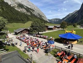 „Blick über das gut besuchte Festgelände des Almkirchtags auf der Eng mit Sitzgarnituren, Almhütten, Verkaufsständen und umliegender Berglandschaft.“