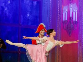 Tänzerin in einem rosafarbenen Ballettkleid führt eine arabeske Position aus, während der als Nussknacker verkleidete Tänzer hinter ihr steht und sie unterstützt.