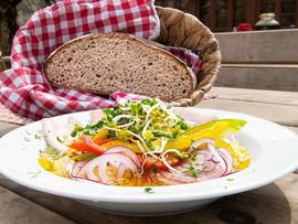 Ein angerichtetes Gericht auf einem weißen Teller mit buntem Gemüse, Sprossen und Zwiebeln. Dahinter liegt eine Scheibe kräftiges Bauernbrot in einem Korb mit rot-weiß karierter Stoffserviette. Das Gericht wird auf einer rustikalen Holzterrasse serviert.