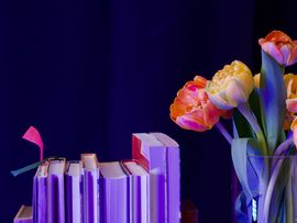 ©M.Kauz Stapel von Büchern neben einer Glasvase mit orange- und rosafarbenen Tulpen vor dunkelblauem Hintergrund.