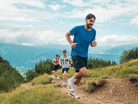 Drei Trailrunner laufen auf einem alpinen Bergweg durch eine grüne Landschaft mit weitem Blick auf die Berge.