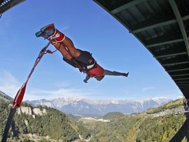 „Bungy-Springer hängt kopfüber an einem Seil unter einer hohen Brücke vor alpiner Berglandschaft.“