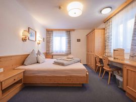 Helles Doppelzimmer der Schlickeralm mit einem großen Holzbett, karierten Textilien, Holzschrank und Schreibtisch. Große Fenster und warme Beleuchtung schaffen eine gemütliche Atmosphäre.