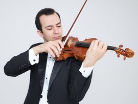 Ein Violinist im schwarzen Anzug spielt eine braune Geige mit konzentriertem Blick vor hellem Hintergrund.