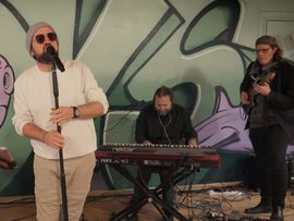 Eine Musikgruppe wo der Sänger eine Runde Brille mit Mütze trägt