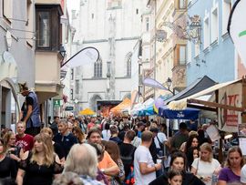 Eine gut besuchte Altstadtgasse mit zahlreichen Marktständen und Besucherinnen und Besuchern. Im Hintergrund ist eine Bühne vor einer Kirche aufgebaut, während die Menschen durch das Festgelände schlendern.