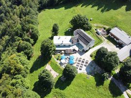 Luftaufnahme des Alpengasthofs St. Magdalena inmitten einer weitläufigen grünen Wiesenlandschaft am Waldrand. Mehrere Gebäude, ein Gastgarten mit Sitzplätzen und Sonnenschirmen sowie Zufahrtswege sind zu sehen.