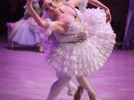 Ballettpaar auf der Bühne: Eine Tänzerin in einem weißen Tutu wird von einem Tänzer in weiß-silbernem Kostüm seitlich gehalten und in einer ausdrucksstarken Pose unterstützt.
