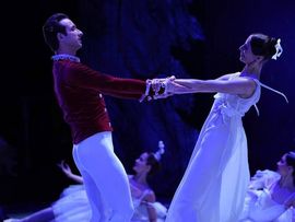Ballettpaar in einer romantischen Szene auf einer blau beleuchteten Bühne. Der Tänzer trägt ein rotes Oberteil und die Tänzerin ein weißes Kleid; beide halten sich an den Händen.