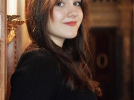 Porträt der Sopranistin Anija Lombard. Sie lehnt an einem Türrahmen und blickt mit einem dezenten Lächeln in die Kamera.