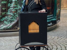 Person mit Schirmmütze steht neben einer historischen Drehorgel auf einem festlich dekorierten Weihnachtsmarkt.