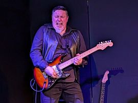Ein Gitarrist der Band Schwarzes Edelweiss spielt mit sichtbarer Begeisterung auf einer Bühne und hält eine E-Gitarre in den Händen.