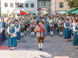 Eine Musikkapelle in traditioneller Tiroler Tracht spielt auf einem Marktplatz. Zahlreiche Besucherinnen und Besucher verfolgen den Auftritt.