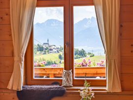 Blick aus dem holzvertäfelten Gastraum durch ein Fenster auf eine alpine Landschaft. Im Vordergrund stehen ein gedeckter Holztisch und eine Vase mit Blumen, während draußen eine Ortschaft und markante Berggipfel zu sehen sind.