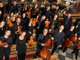 Ein Orchester mit Streich-, Holz- und Blechblasinstrumenten posiert in einer Kirche für ein Gruppenfoto. Die Musikerinnen und Musiker halten ihre Instrumente in den Händen und tragen schwarze Konzertkleidung.