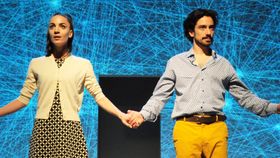 Mann und Frau vor abstraktem Bühnenhintergrund blicken händehaltend Richtung Publikum